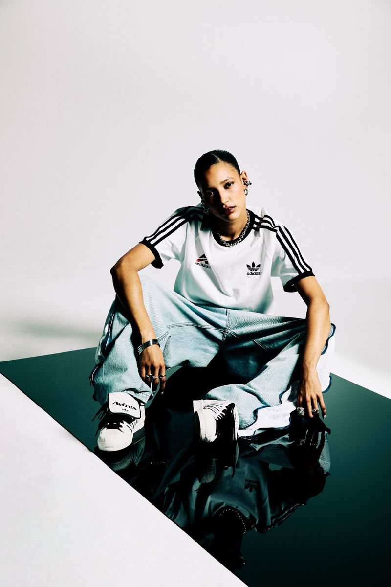 Young woman in Adidas gear sits on reflective surface. She wears baggy jeans, a striped tee, sneakers, jewelry, confident pose, indoor setting.