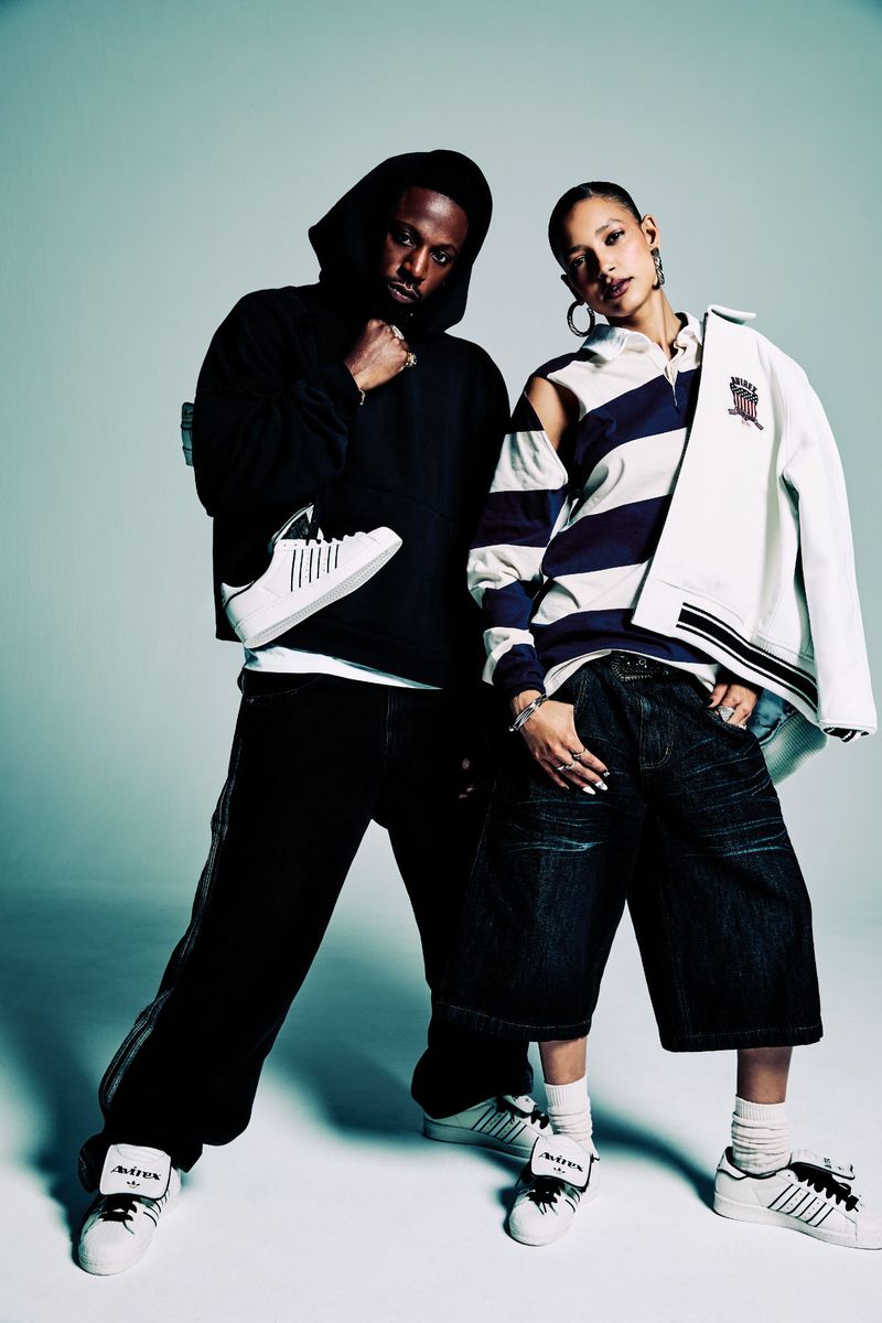 Two people in a fashion shot: an adult male in a black hoodie holding a white shoe and an adult female in oversized denim shorts.