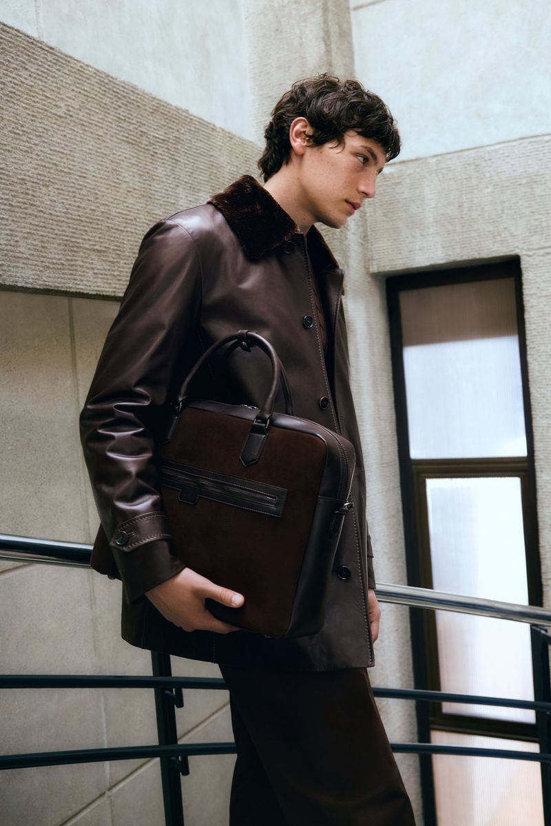 Man in leather coat and pants carrying a leather briefcase. Indoor shot in a stairwell setting. Fashion editorial photography.