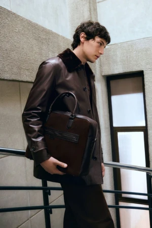 Man in leather coat and pants carrying a leather briefcase. Indoor shot in a stairwell setting. Fashion editorial photography.