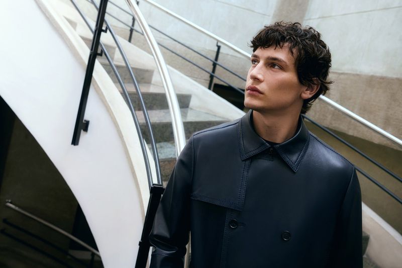 Adult male poses on a staircase wearing a stylish blue-grey leather trench coat in an indoor environment under soft light.