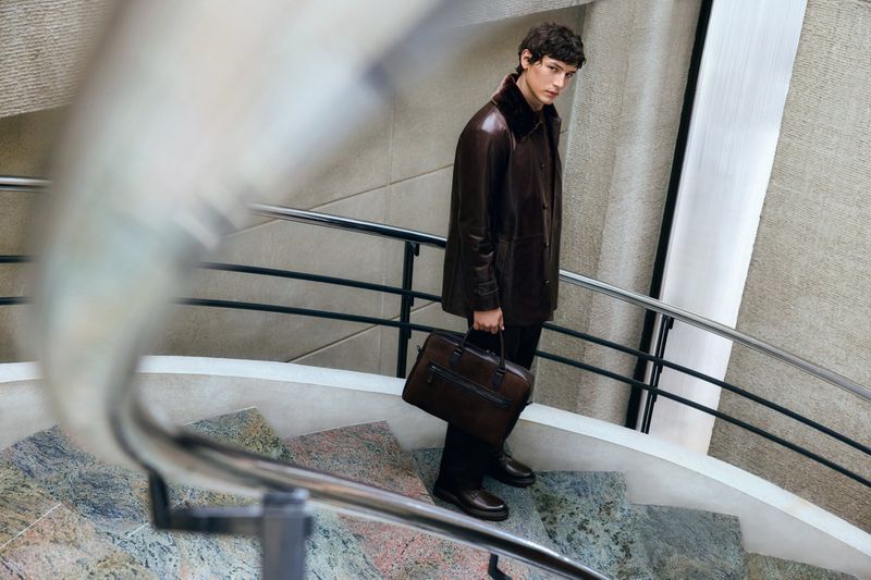 Man in leather coat walks down marble stairs holding a brown briefcase. Modern architecture with cool grey hues and steel handrails.