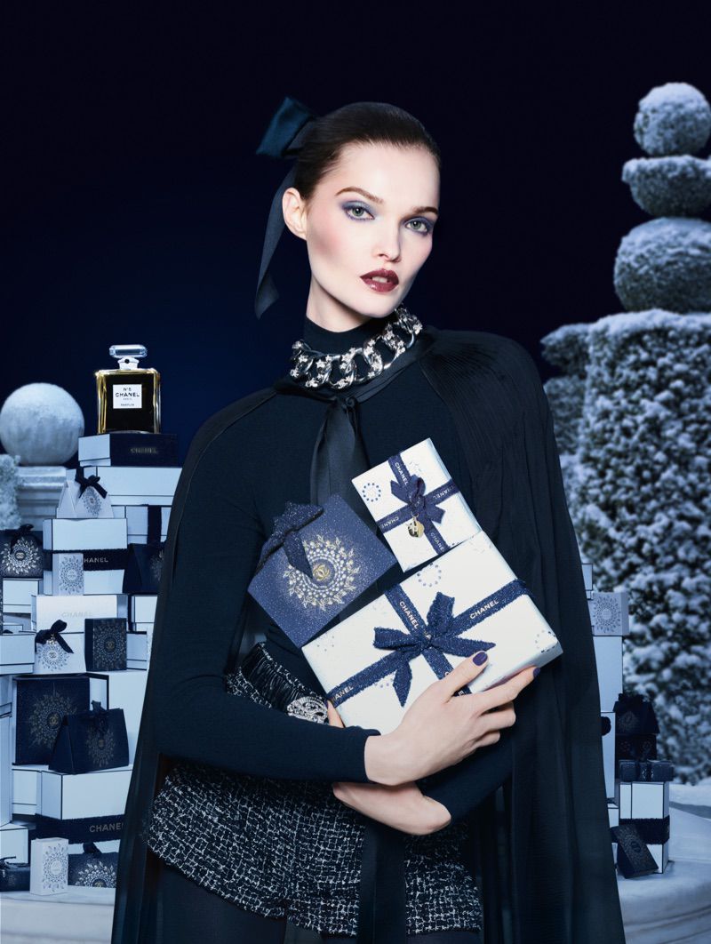 Elegant woman in a black dress holds gift boxes near perfume and decorative snow-covered trees against a dark blue backdrop.