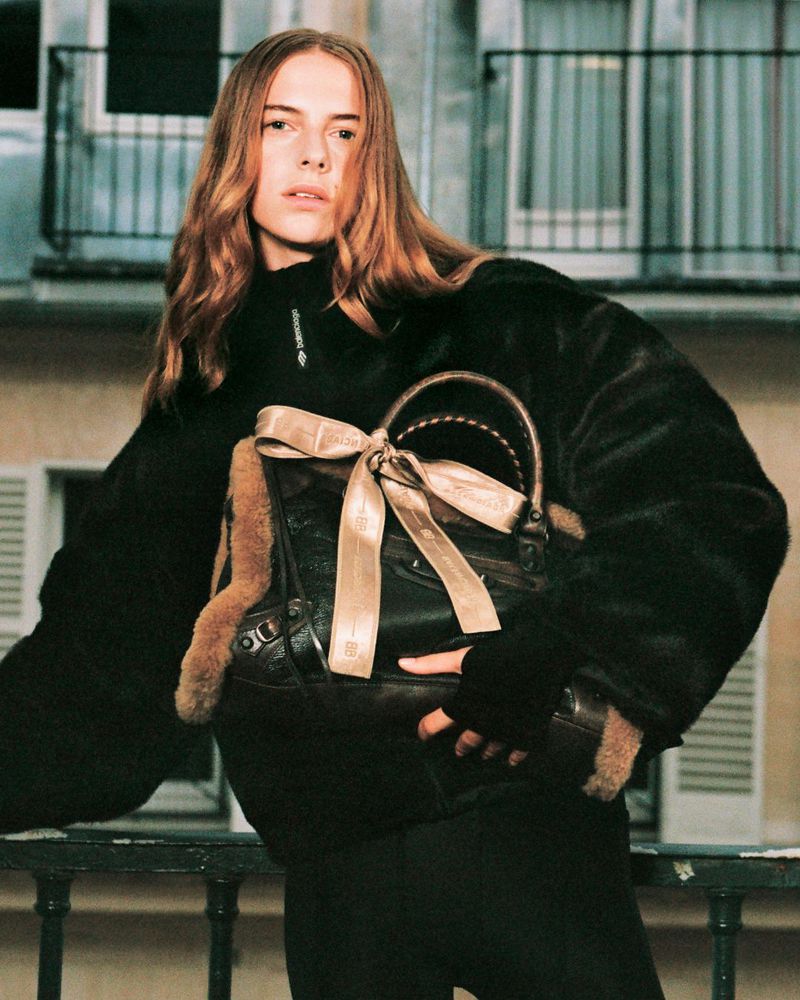 Woman in a black fur coat standing on a balcony with a purse with a ribbon in front of a building with windows and shutters.