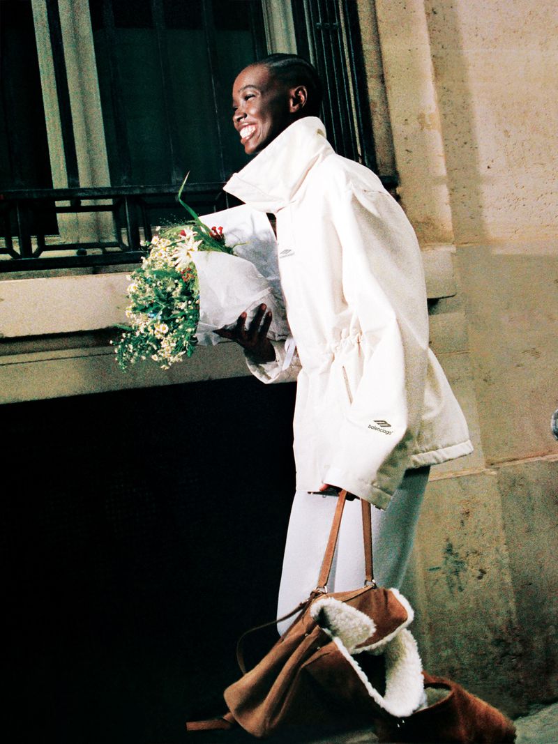 A smiling woman with short dark hair wearing a white jacket walks out of a building holding a bouquet of white and green flowers.