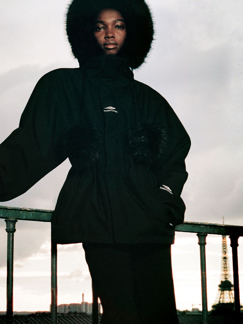 Fashion editorial shot of a woman wearing a black hooded parka jacket with fur trim standing in front of Paris cityscape with Eiffel Tower.