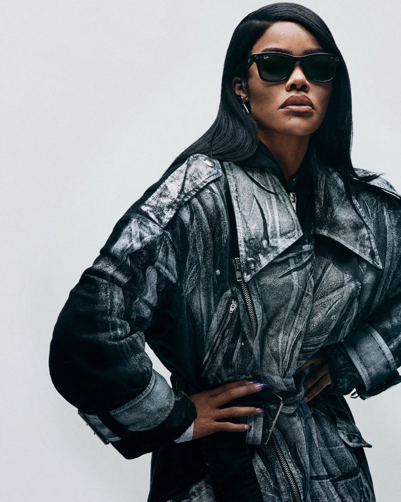 Adult female with long black hair poses in a black and white patterned jacket and sunglasses against a neutral background in a studio shot.