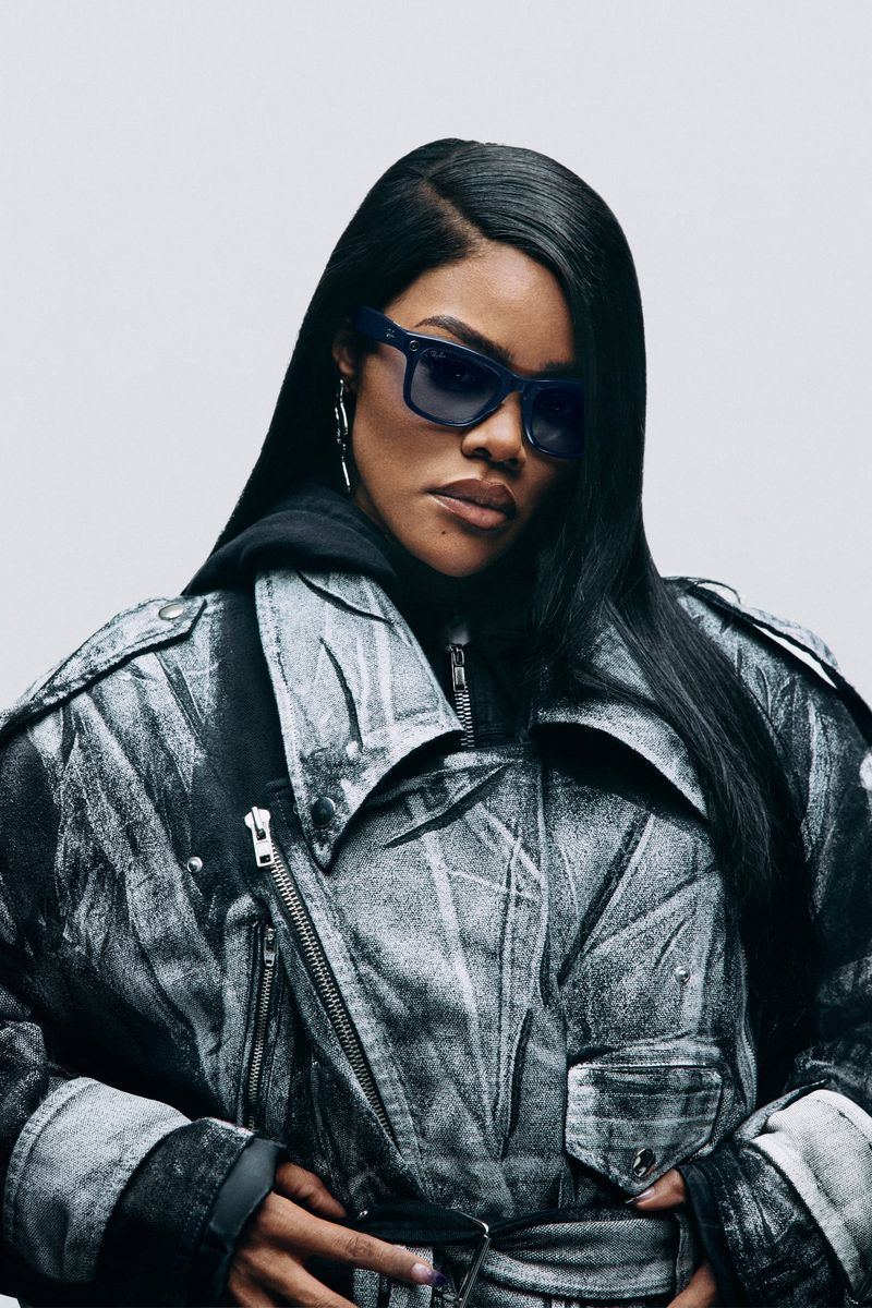 Adult woman in sunglasses and grey moto jacket, indoor studio portrait against a neutral background, long dark hair and casual style.