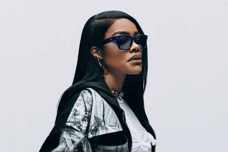 Portrait of an adult female with long black hair, wearing sunglasses, a white top, and a black and white jacket against a white backdrop.