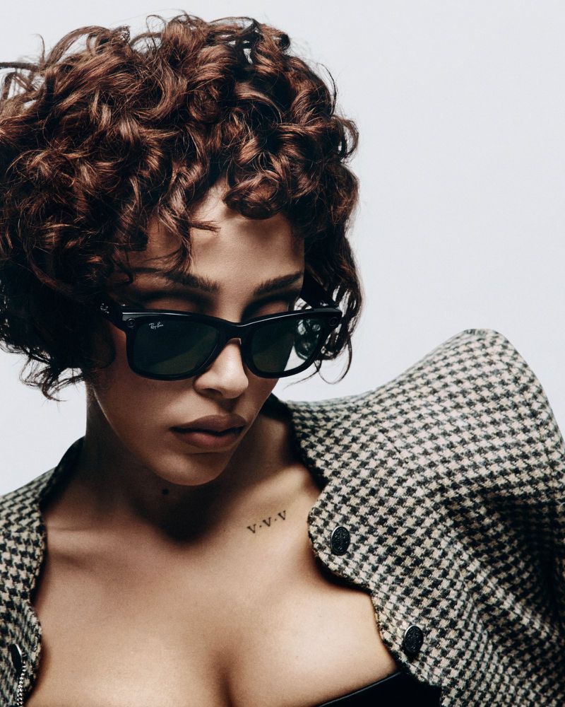 Portrait of a curly haired woman wearing sunglasses and a patterned jacket against a white background in a studio setting.