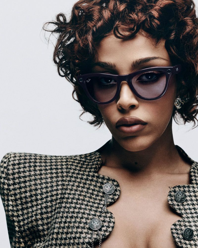 Portrait of a woman with auburn curly hair, wearing a patterned jacket, statement earrings, and purple sunglasses in a studio setting.