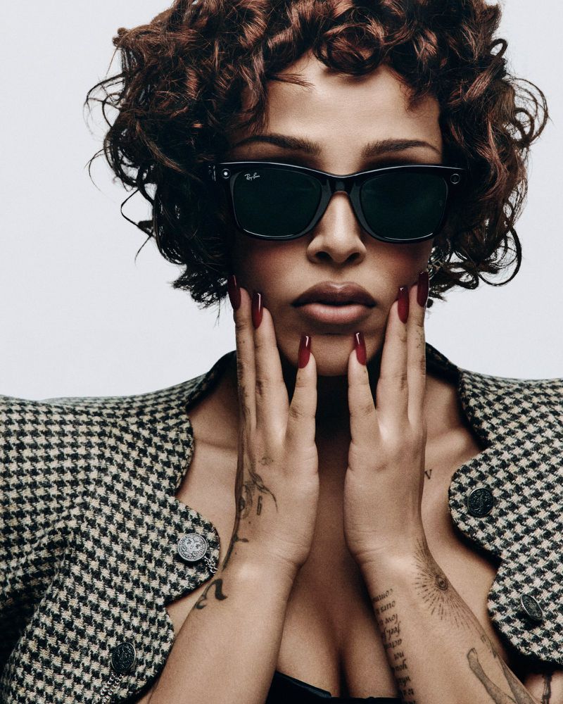 Portrait of a woman with an afro and sunglasses, wearing a patterned jacket, hands near her face, posing indoors against a bright backdrop.
