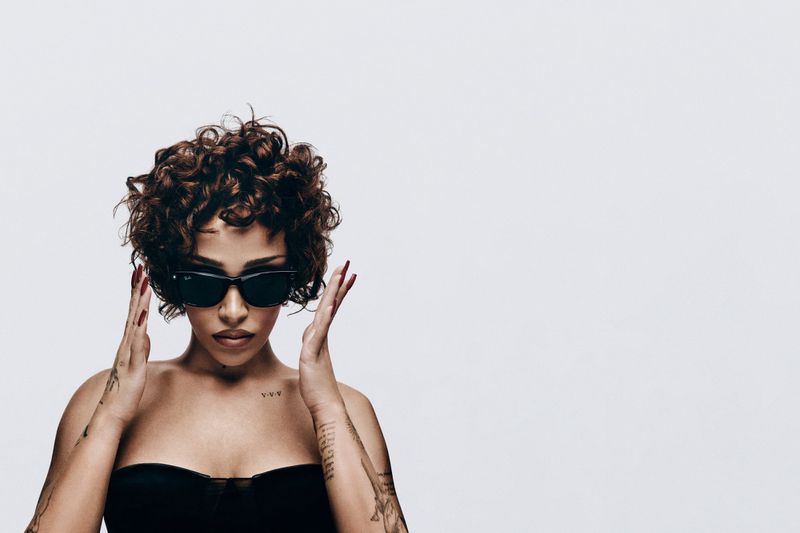Portrait of woman with curly hair, sunglasses, and bare shoulders in a studio, with a tattoo on her arm, set against a white backdrop.