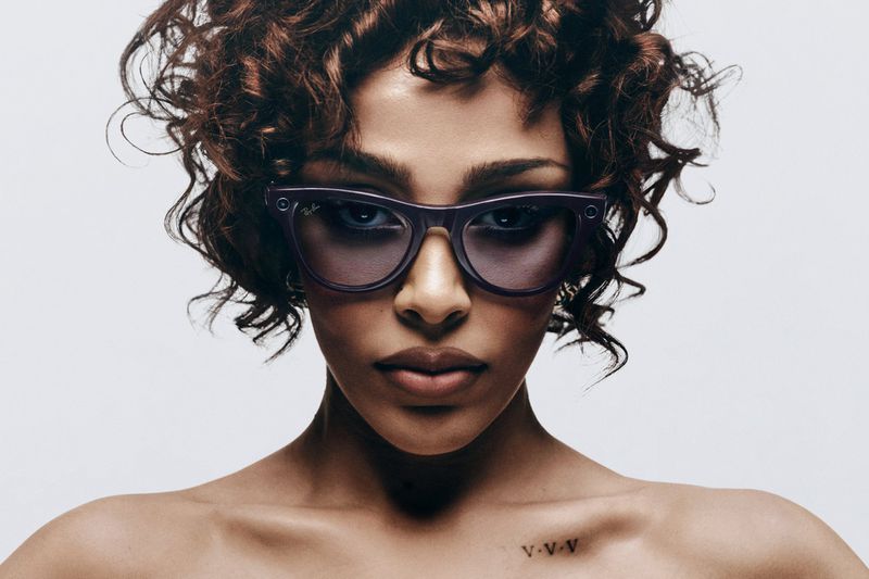 Close-up portrait of a brunette woman with curly hair, wearing purple glasses against a clean, light background in a studio setting.