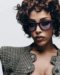 Doja Cat wearing purple Ray-Ban Meta smart glasses and a houndstooth patterned jacket against a white background