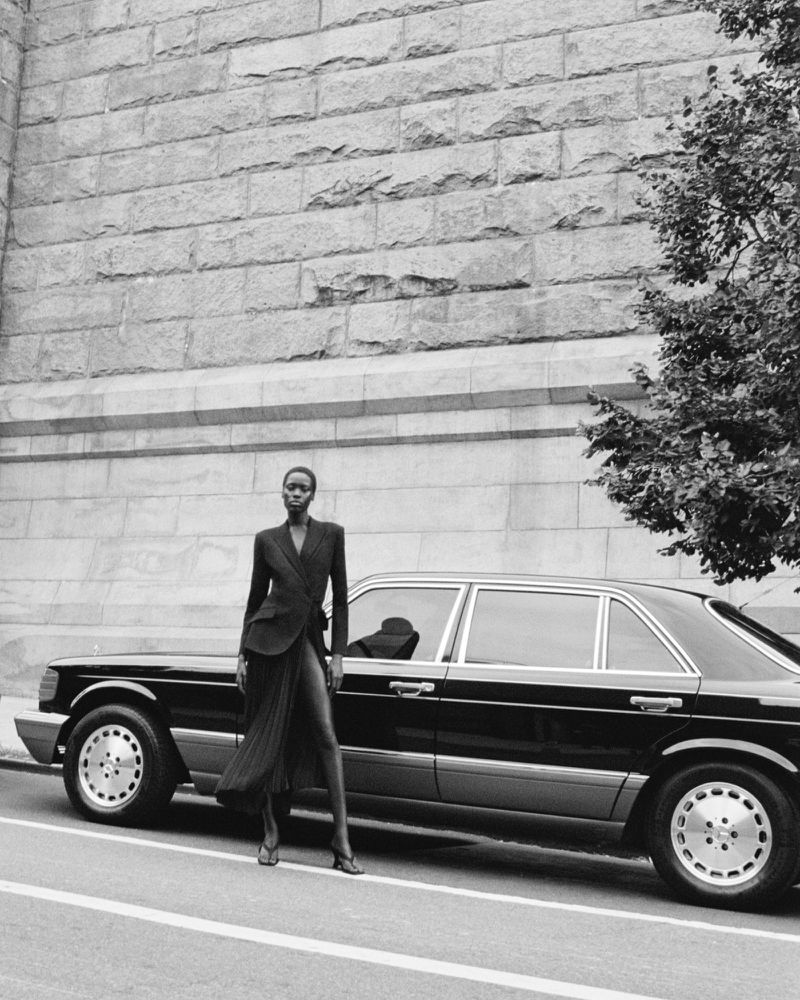 A slender woman poses next to a vintage automobile on an urban street wearing an elegant wrap blazer and flowing skirt in a grayscale image.