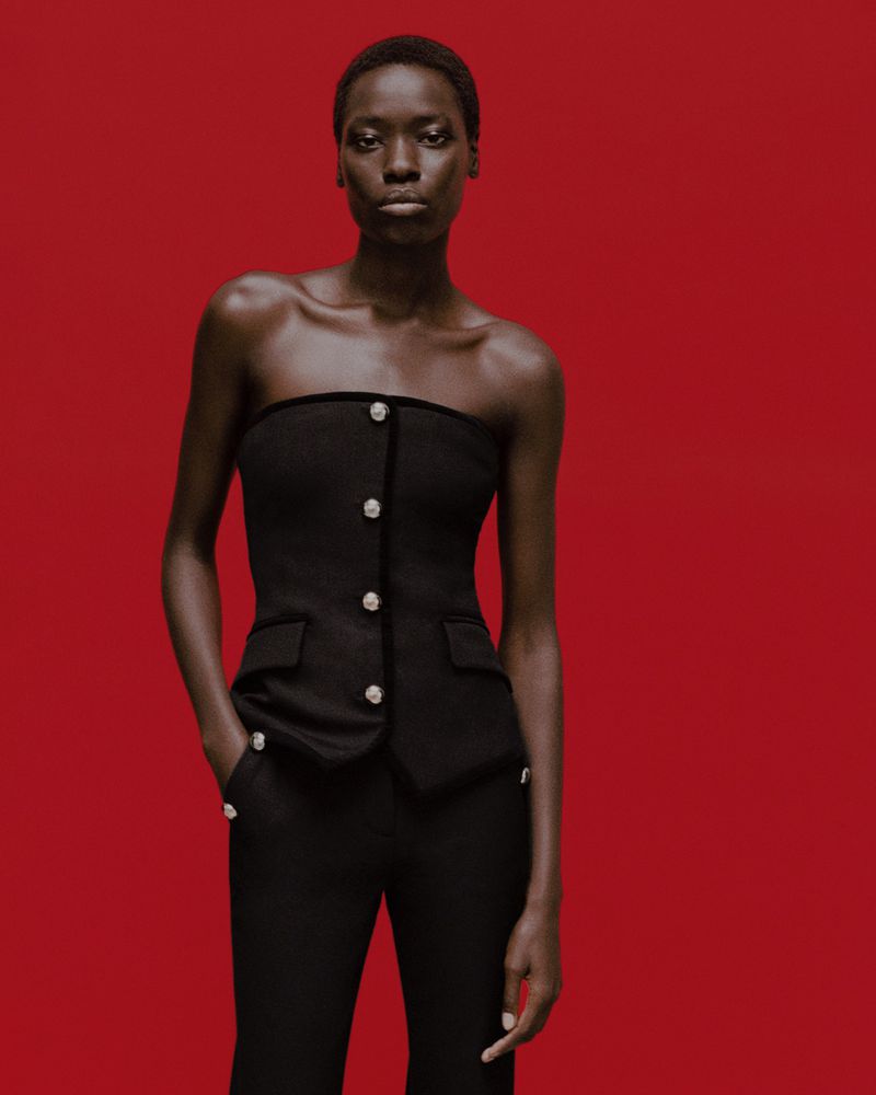 Fashionable dark-skinned woman in a strapless black top and black pants posing against a solid red background in a studio setting.