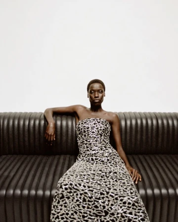 Model Awar Odhiang seated on a dark fluted sofa in a strapless metallic patterned gown for Simkhai Holiday 2025.