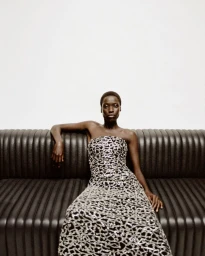 Model Awar Odhiang seated on a dark fluted sofa in a strapless metallic patterned gown for Simkhai Holiday 2025.