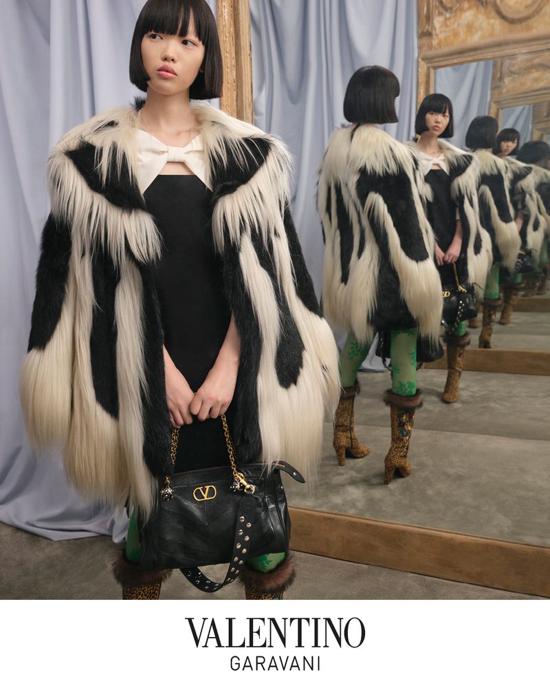 Elegant woman in a black dress and faux fur coat holding a black Valentino Garavani purse in front of a mirror.