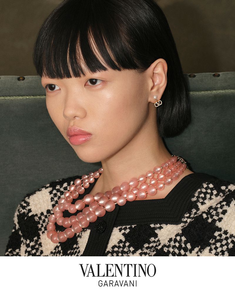 Close-up of an Asian female model wearing a pink pearl necklace and designer earrings, showcasing fashion and beauty in an indoor setting.