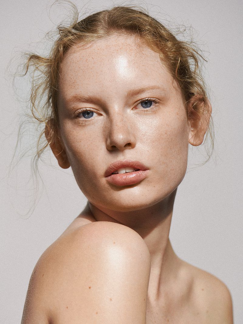 Close-up studio portrait of a young adult woman with freckles and blue eyes looking at the camera with a neutral expression.