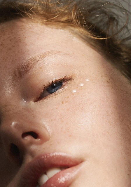 Close-up portrait of a fair-skinned woman with freckles, blue eyes, and light red hair, featuring three small cosmetic cream drops under her eye.
