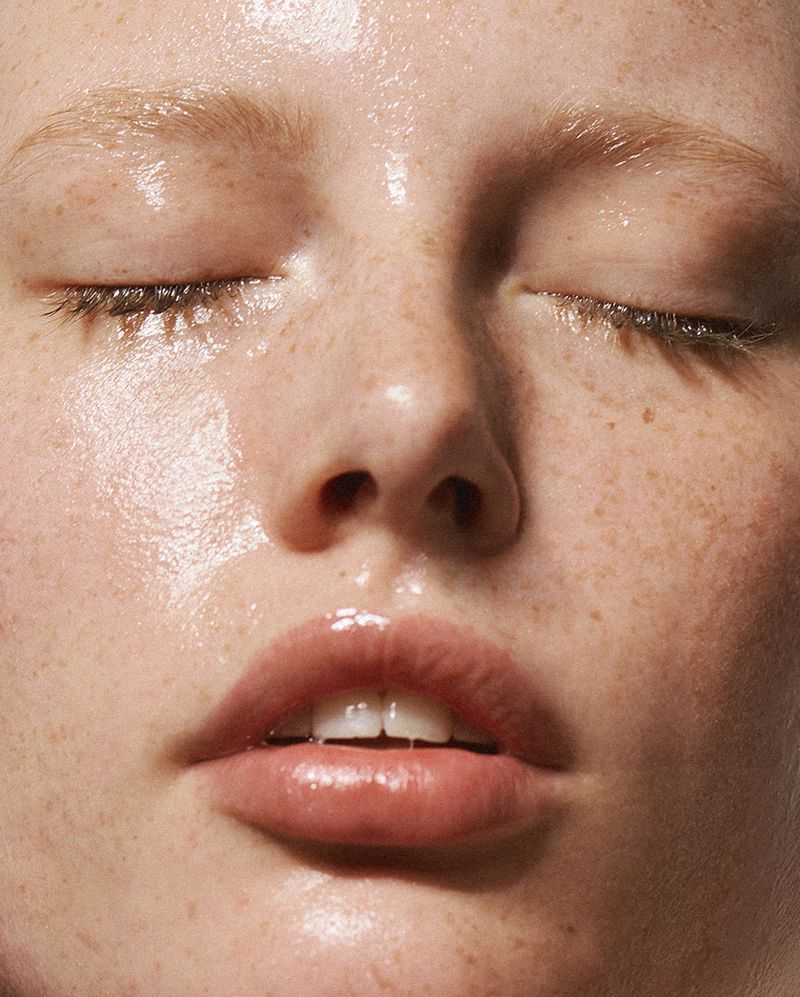 Close-up portrait of a woman with freckles and closed eyes, glossy skin, and pink lips, showcasing beauty and skincare products in soft lighting.