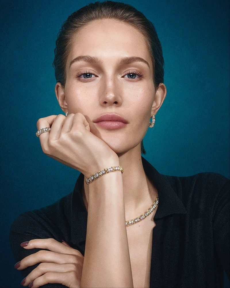 Close-up of an adult woman with light skin, wearing jewelry and a black top against a teal background in a studio shot.