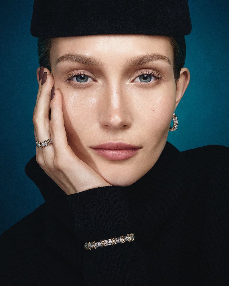 Portrait of a fair-skinned woman with light eyes, wearing a black hat and turtleneck, accented with jewelry, against a teal backdrop.