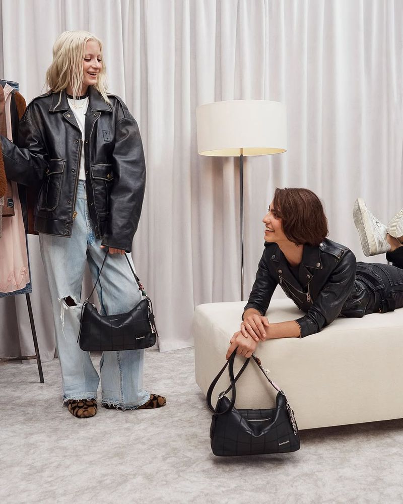 Two women pose indoors wearing leather jackets and holding handbags. One woman is reclining on a white bench in a studio-like space.