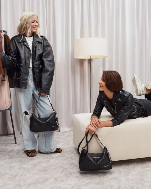 Two women pose indoors wearing leather jackets and holding handbags. One woman is reclining on a white bench in a studio-like space.