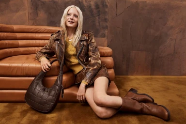 Model with blonde hair wearing a leather jacket and holding a dark brown handbag while sitting on a brown sofa