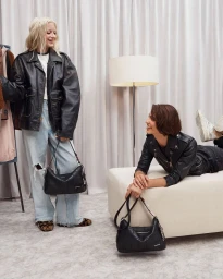 Two models posing with black leather handbags in a studio set with white draped curtains and a floor lamp