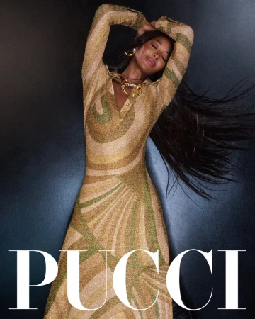Elegant adult female with long hair poses in a patterned gold dress and jewelry against a dark background for a fashion editorial.