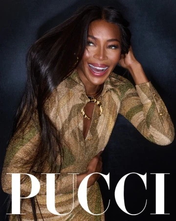 Elegant portrait of Naomi Campbell in a patterned gold dress, smiling with her long black hair cascading down against a dark backdrop.