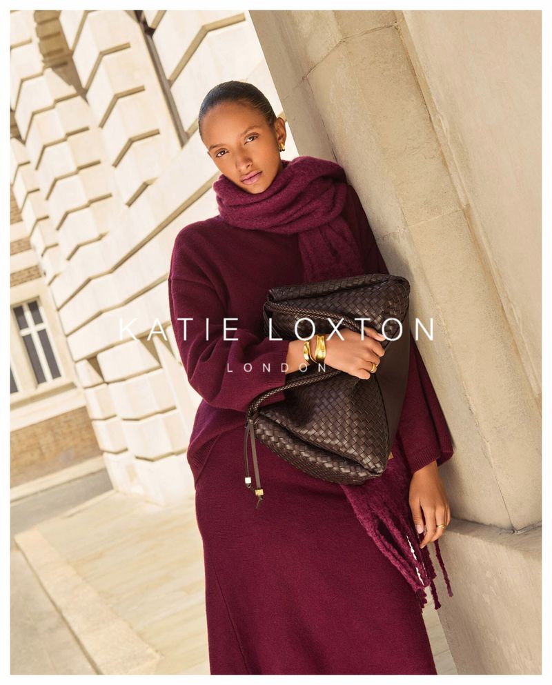 Brunette woman poses in a burgundy knit dress, scarf and carries woven handbag against a neutral building backdrop for fashion ad.