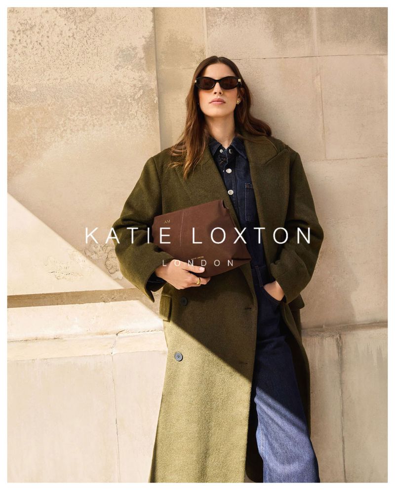 A stylish adult woman in an olive coat and sunglasses poses while holding a brown clutch bag against a textured wall.