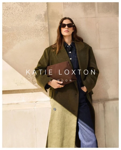 A stylish adult woman in an olive coat and sunglasses poses while holding a brown clutch bag against a textured wall.