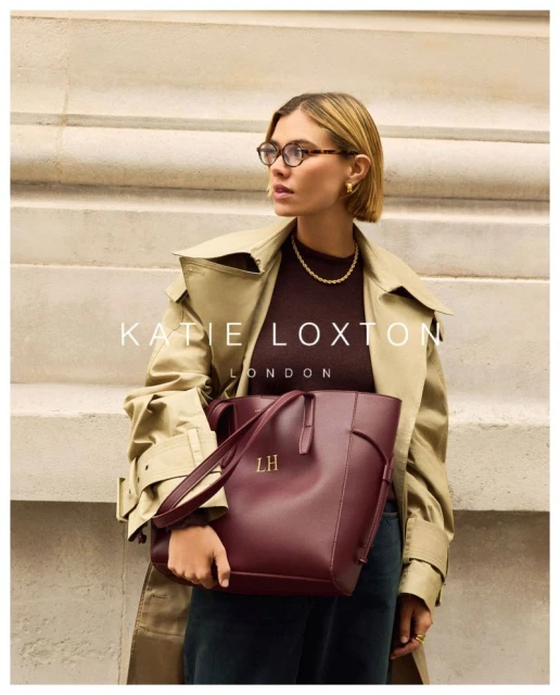 Adult woman with bobbed hair poses in a tan trench coat holding a burgundy Katie Loxton tote bag in an outdoor setting.