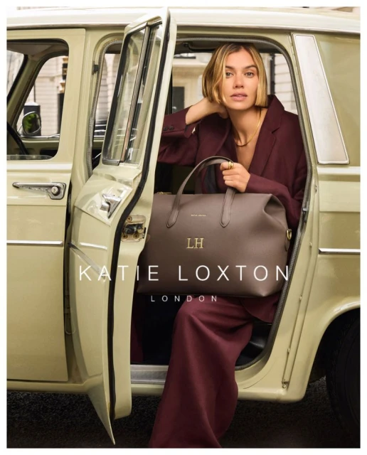 Blonde woman in maroon suit poses in cream-colored car with large brown handbag from Katie Loxton London, creating a fashion moment.