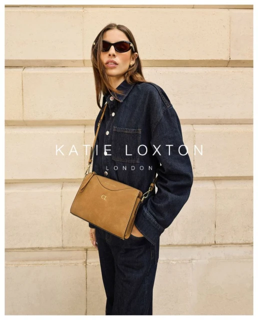 Woman in denim jacket and jeans poses with brown leather shoulder bag against a neutral wall, showcasing fashion and style.