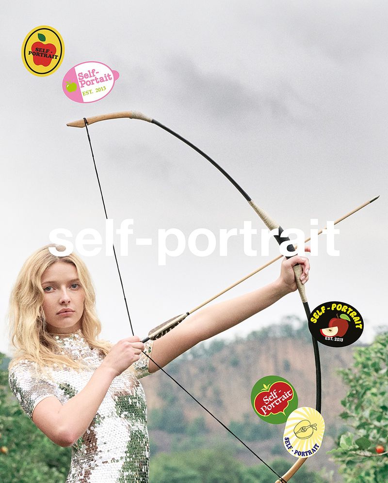 Woman in a sparkly dress poses with a bow and arrow in an orchard with trees and a cloudy sky. Fashion editorial.
