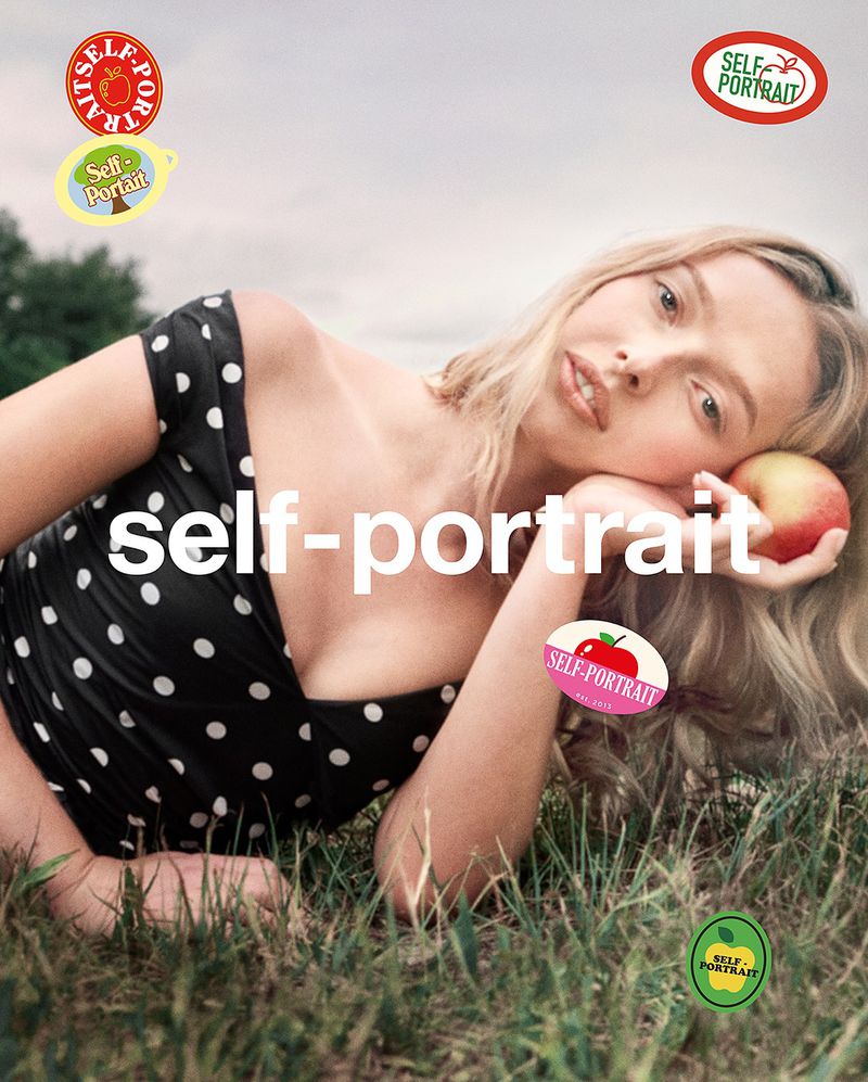 Blonde woman in polka dot dress lies in grass holding apple, posing for a self-portrait fashion advertisement against a cloudy sky.