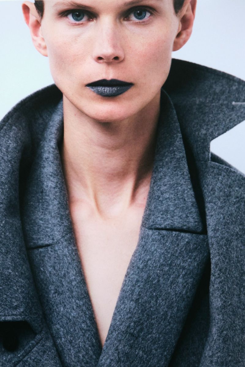 Close-up of a fair-skinned young adult with dark lipstick, and grey wool coat on a neutral background in a studio-like setting.