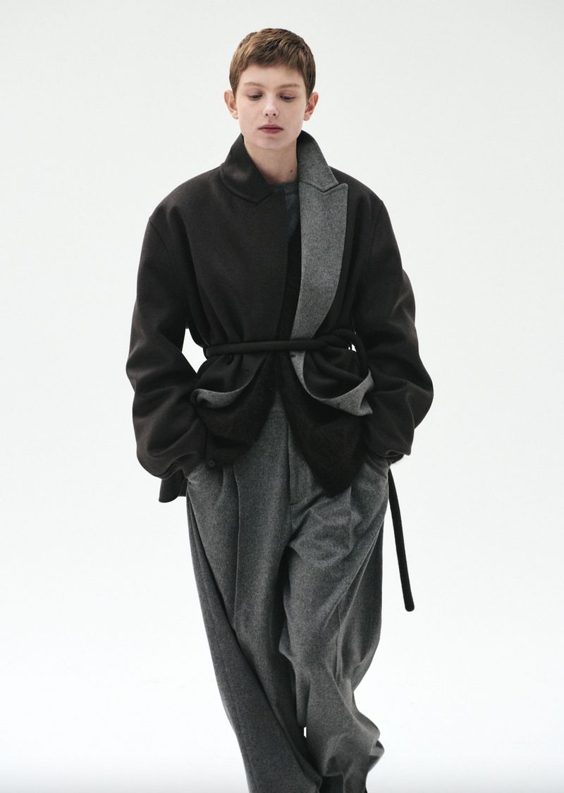 A woman in an overcoat with a rope belt and wide-leg trousers stands in a full shot in front of a white backdrop in a fashion editorial.