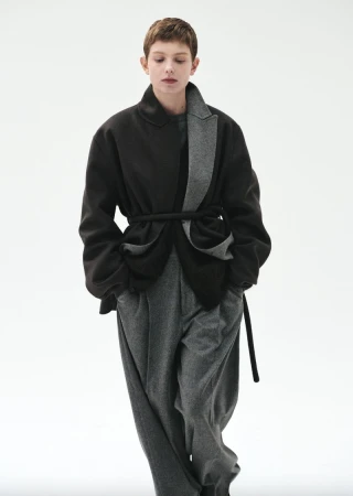 A woman in an overcoat with a rope belt and wide-leg trousers stands in a full shot in front of a white backdrop in a fashion editorial.