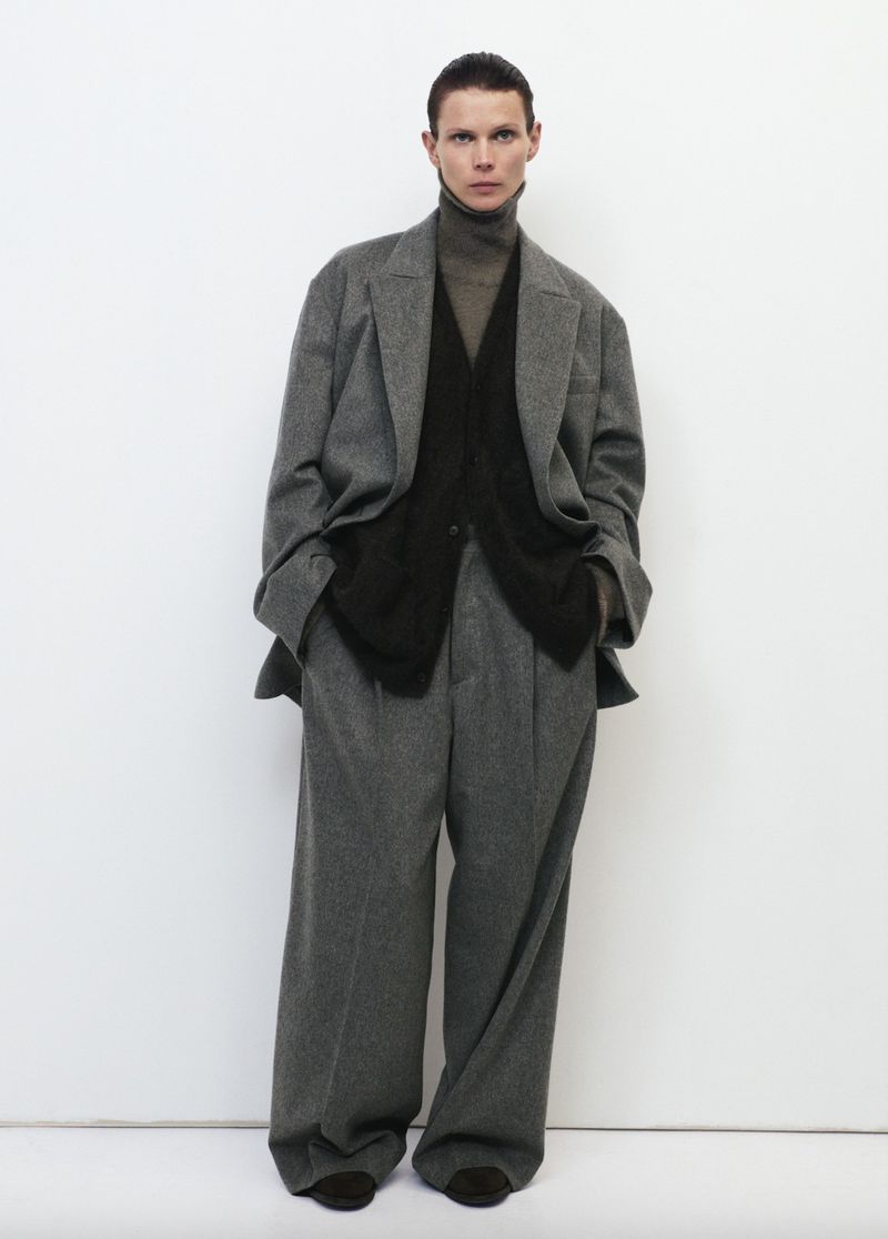 A person stands against a white wall modeling a grey wool suit with wide-legged pants, a black vest, and a brown turtleneck sweater.