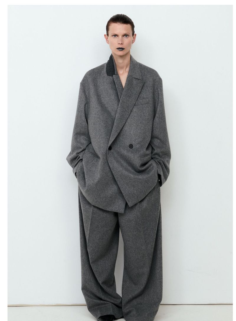 An androgynous person stands in a studio wearing an oversized grey wool suit, showcasing a modern and edgy fashion statement.