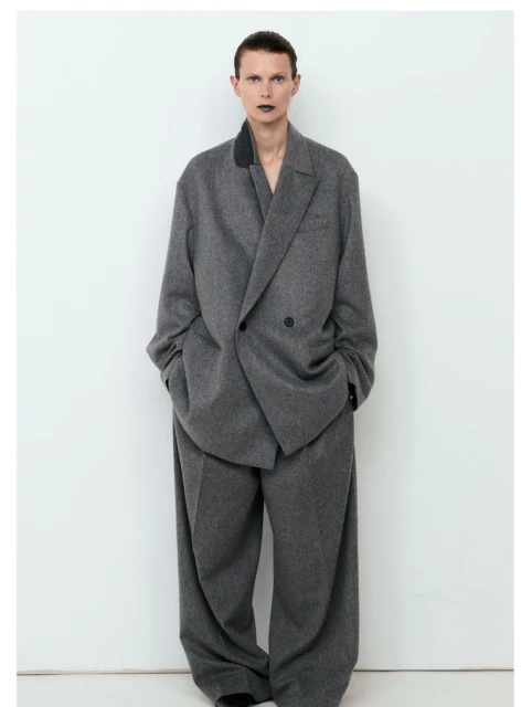 An androgynous person stands in a studio wearing an oversized grey wool suit, showcasing a modern and edgy fashion statement.